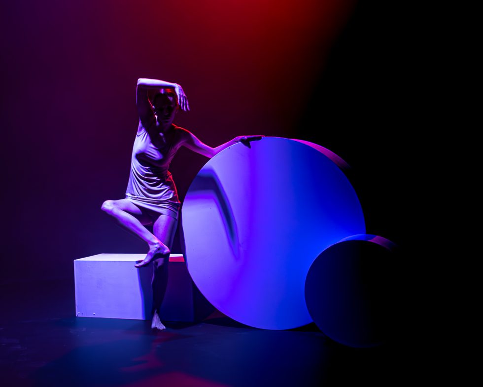 “Elisa, bailarina contemporánea posando entre formas geométricas iluminadas en tonos rojos, violetas y azules durante una sesión fotográfica artística.”