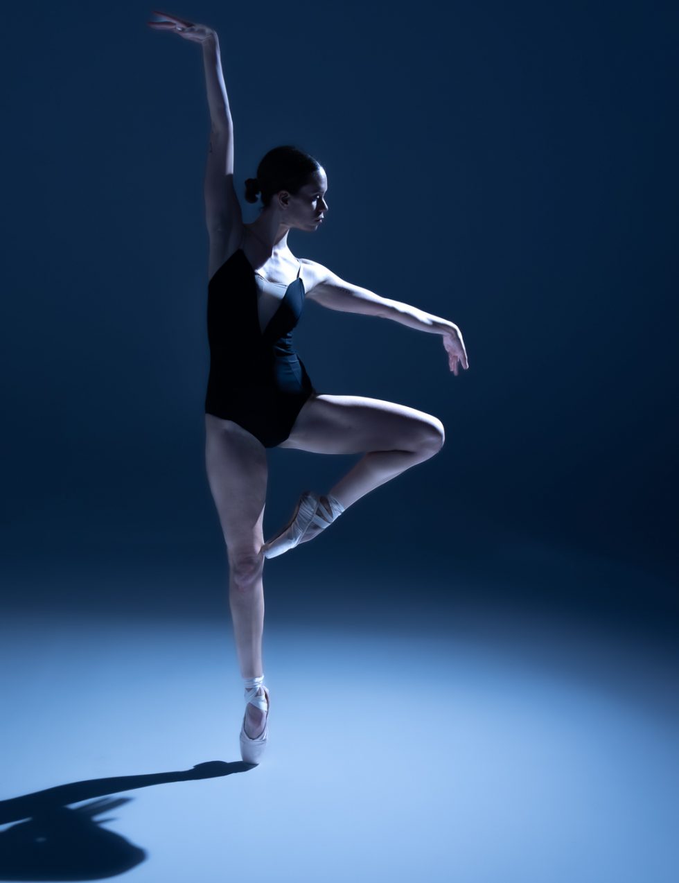 “Bailarina Elisa ejecutando poses de danza clásica y contemporánea sobre punta en estudio, iluminada en tonos azules.”