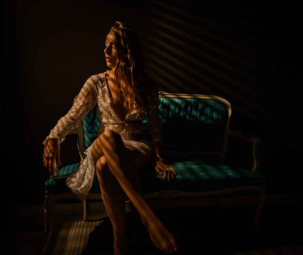 Mujer sentada en un sofá vintage iluminada por luz cálida a través de persianas, con un vestido de encaje.