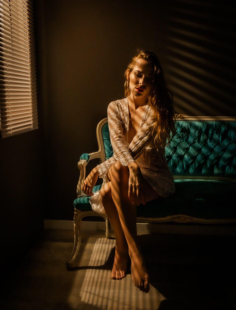 Mujer sentada en un sofá vintage iluminada por luz cálida a través de persianas, con un vestido de encaje.