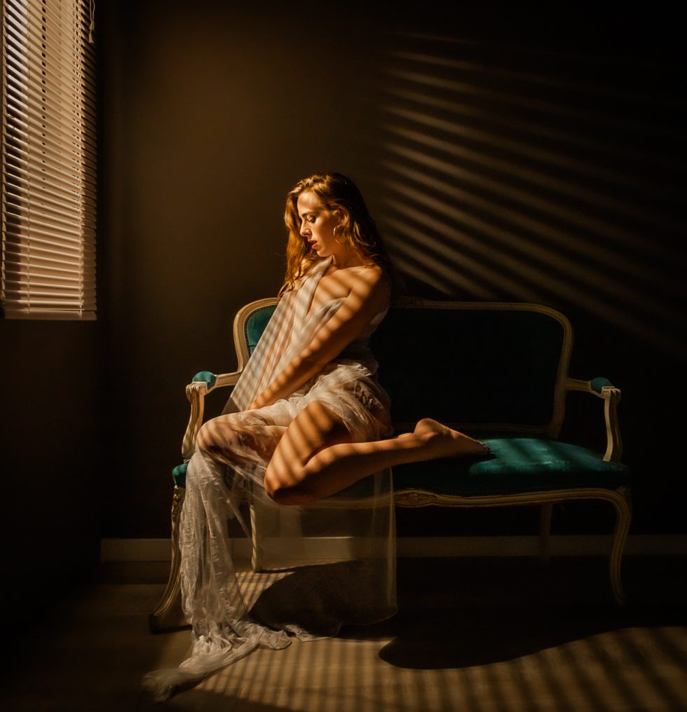 Mujer sentada en un sofá vintage iluminada por luz cálida a través de persianas, con un vestido de encaje.