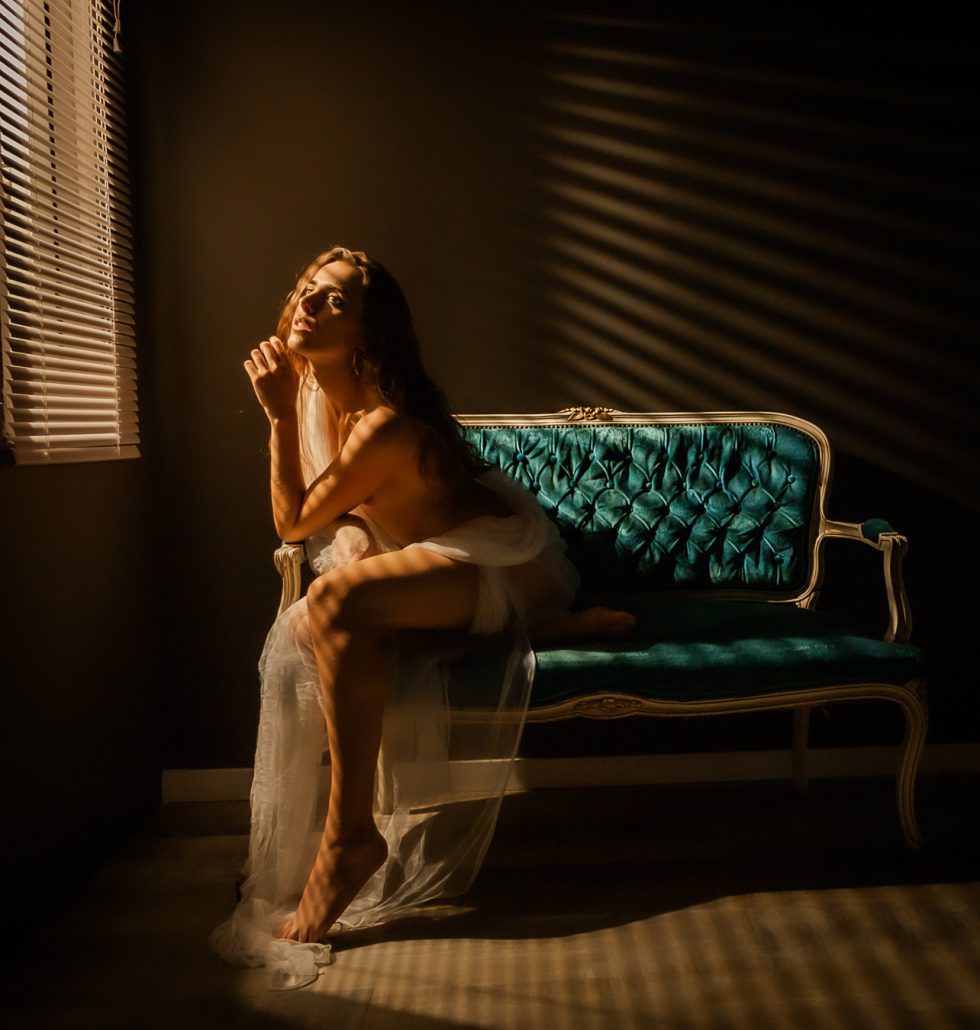 Mujer sentada en un sofá vintage iluminada por luz cálida a través de persianas, con un vestido de encaje.