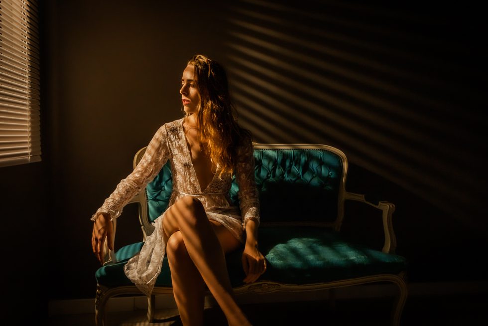 Mujer sentada en un sofá vintage iluminada por luz cálida a través de persianas, con un vestido de encaje.