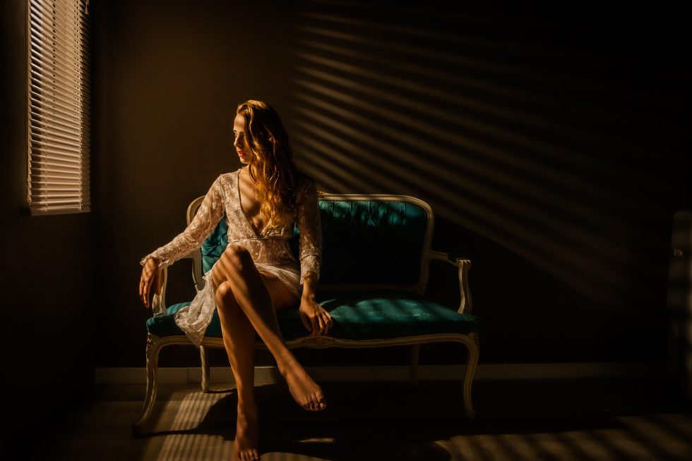 Mujer sentada en un sofá vintage iluminada por luz cálida a través de persianas, con un vestido de encaje.