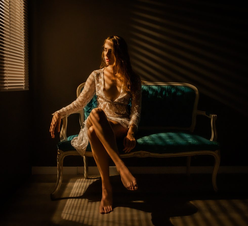 Mujer sentada en un sofá vintage iluminada por luz cálida a través de persianas, con un vestido de encaje.