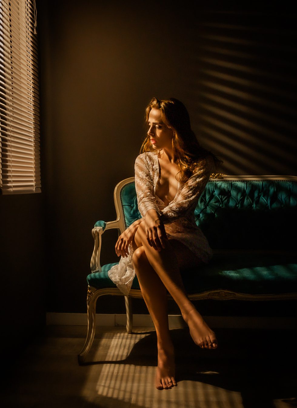 Mujer sentada en un sofá vintage iluminada por luz cálida a través de persianas, con un vestido de encaje.