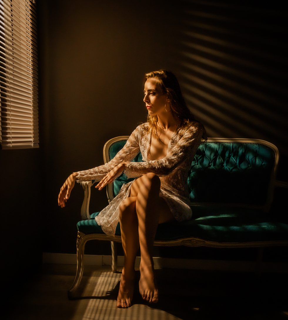 Mujer sentada en un sofá vintage iluminada por luz cálida a través de persianas, con un vestido de encaje.