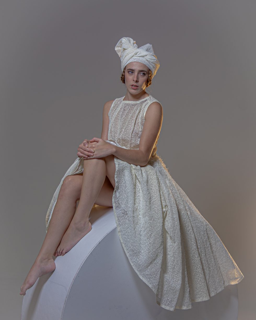 Mujer joven con vestido blanco texturizado y turbante, posando con gestos expresivos sobre una plataforma circular, en un ambiente de estudio con iluminación artística.