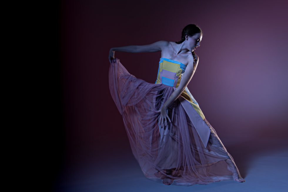 Mujer joven con vestido blanco texturizado y turbante, posando con gestos expresivos sobre una plataforma circular, en un ambiente de estudio con iluminación artística.
