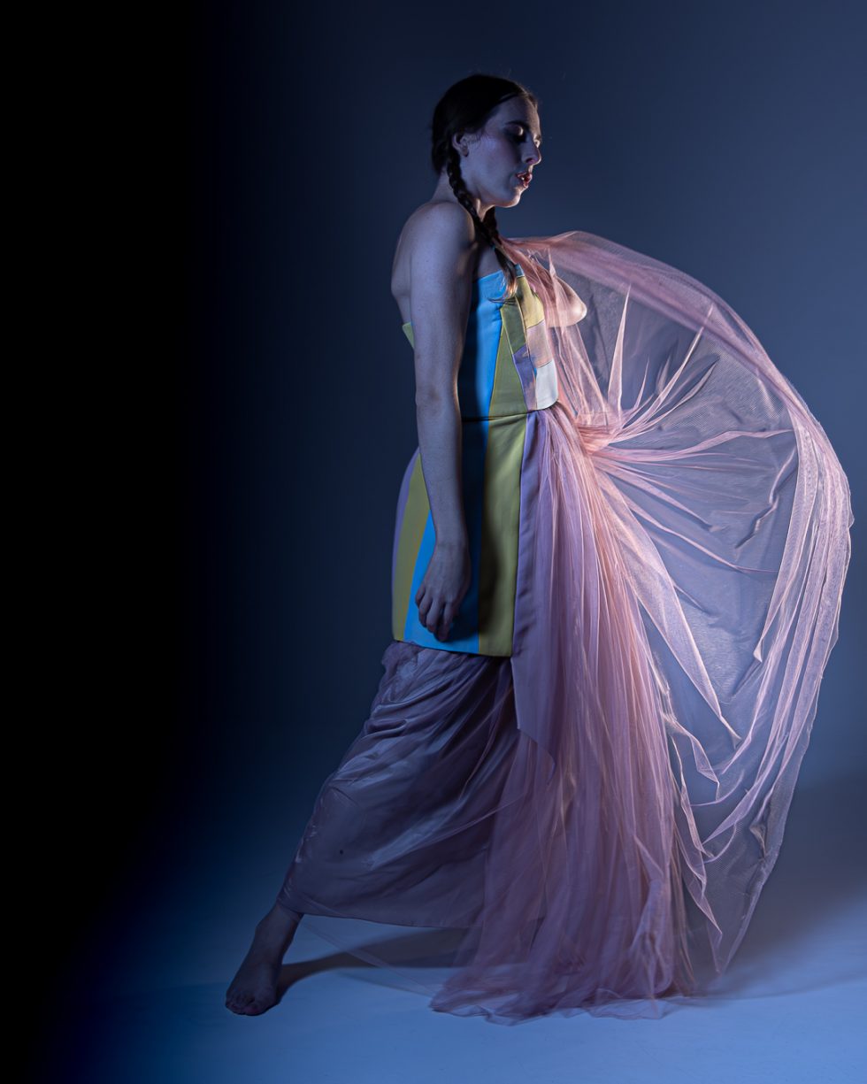 Mujer joven con vestido blanco texturizado y turbante, posando con gestos expresivos sobre una plataforma circular, en un ambiente de estudio con iluminación artística.