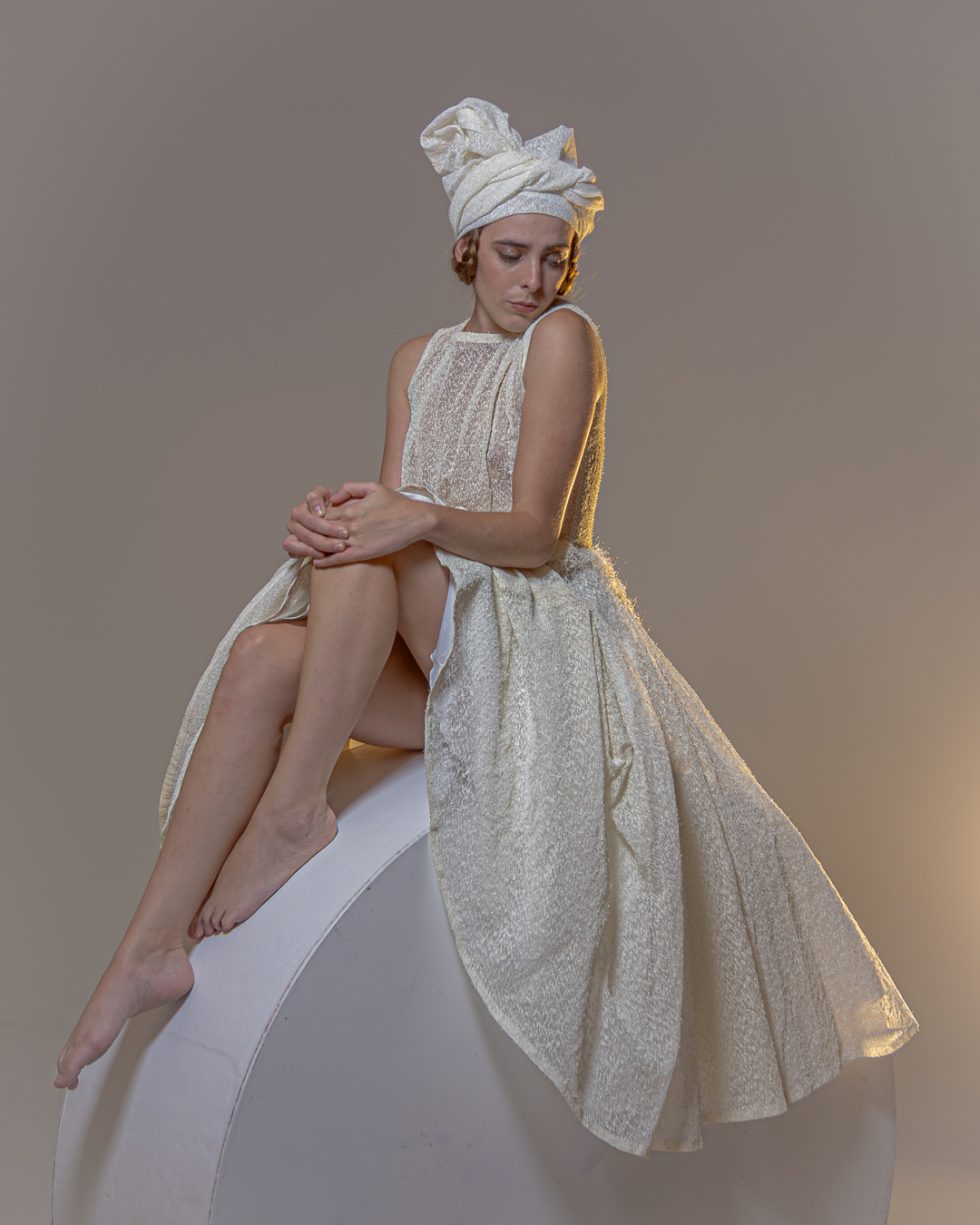 Mujer joven con vestido blanco texturizado y turbante, posando con gestos expresivos sobre una plataforma circular, en un ambiente de estudio con iluminación artística.