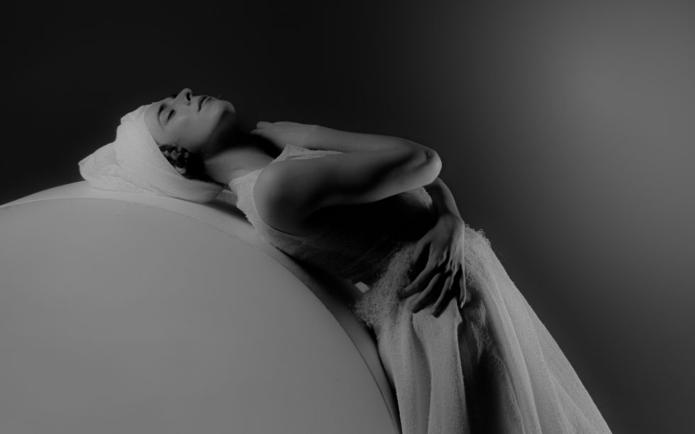 Mujer joven con vestido blanco texturizado y turbante, posando con gestos expresivos sobre una plataforma circular, en un ambiente de estudio con iluminación artística.