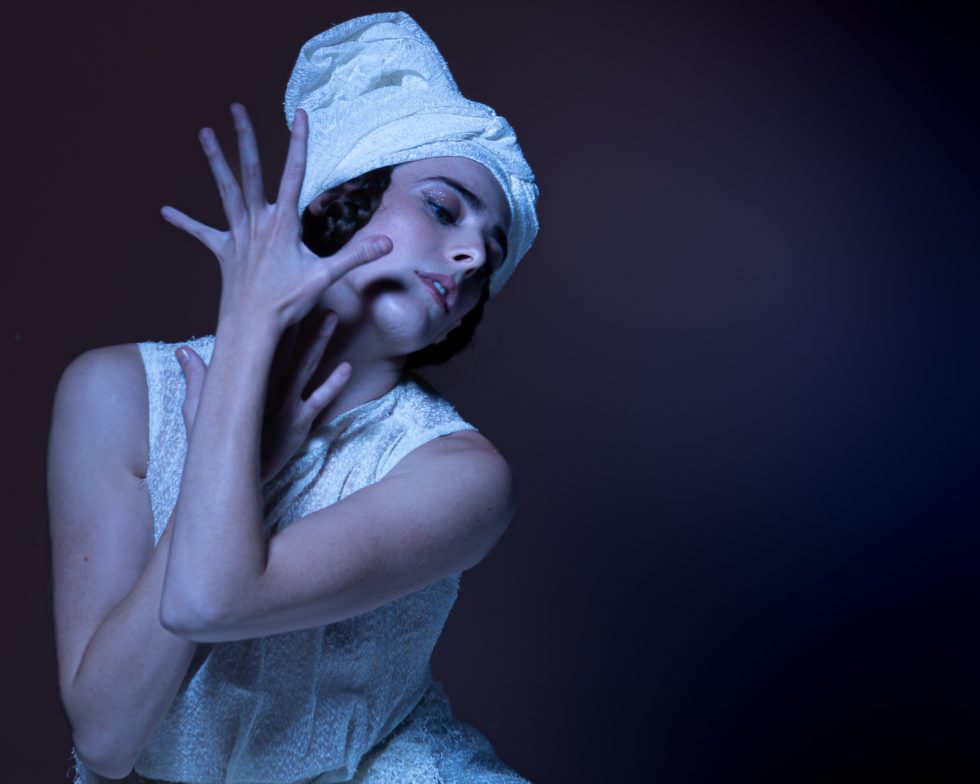 Mujer joven con vestido blanco texturizado y turbante, posando con gestos expresivos sobre una plataforma circular, en un ambiente de estudio con iluminación artística.