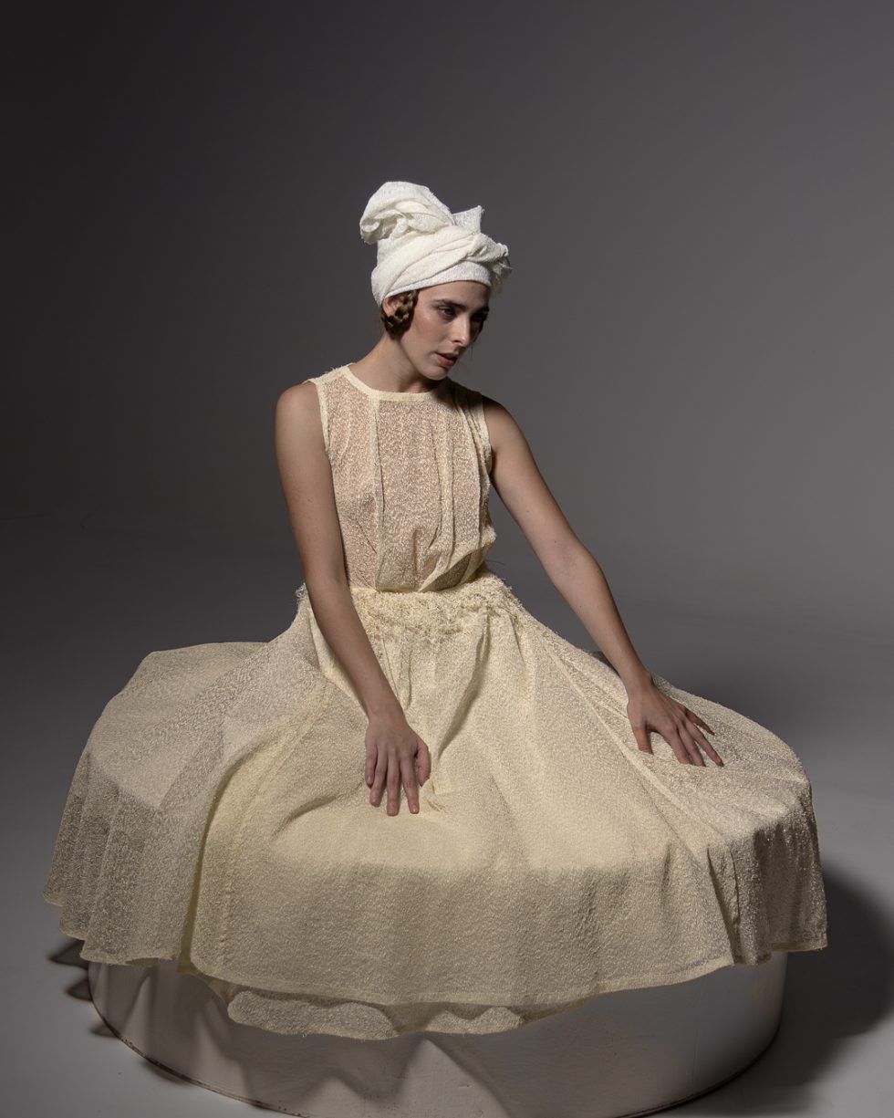 Mujer joven con vestido blanco texturizado y turbante, posando con gestos expresivos sobre una plataforma circular, en un ambiente de estudio con iluminación artística.