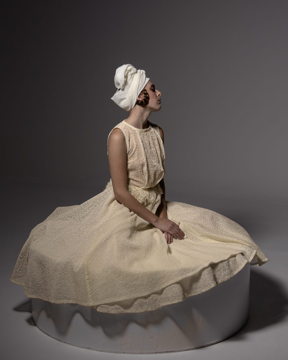 Mujer joven con vestido blanco texturizado y turbante, posando con gestos expresivos sobre una plataforma circular, en un ambiente de estudio con iluminación artística.
