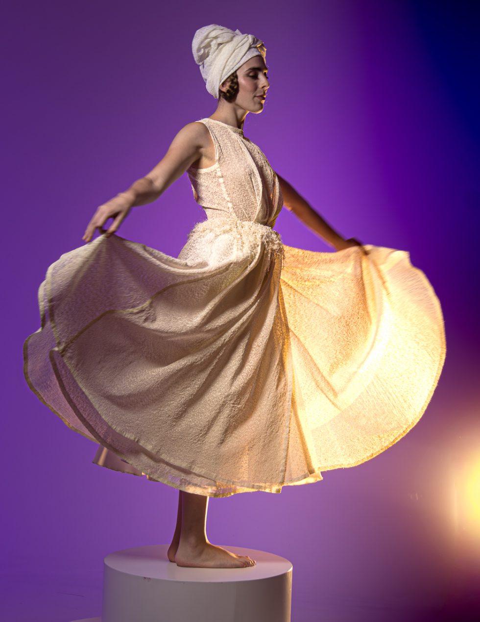 Mujer joven con vestido blanco texturizado y turbante, posando con gestos expresivos sobre una plataforma circular, en un ambiente de estudio con iluminación artística.