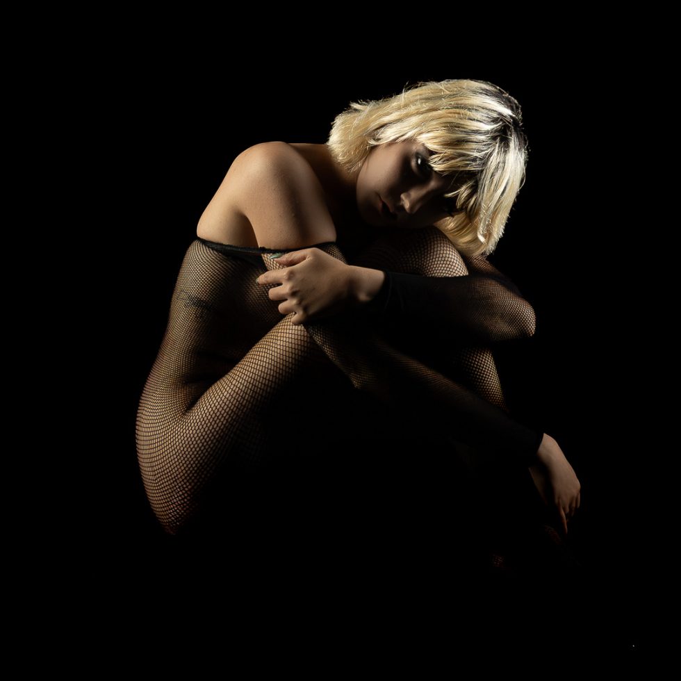Serie de retratos artísticos en clave baja de modelo femenina en estudio, fondo negro, poses introspectivas y sensuales, iluminación dramática y vestuario de malla y transparencias.