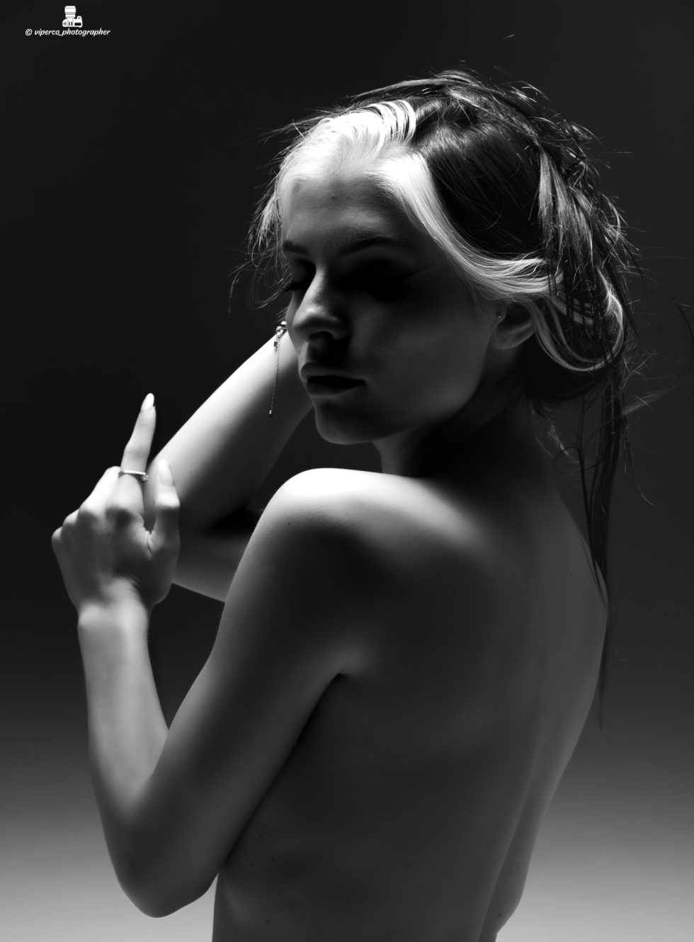 “Mujer arrodillada en un set de fotografía en blanco y negro, con el torso desnudo, cabeza inclinada hacia atrás y brazos abiertos, iluminada por un haz de luz circular que resalta su silueta.”