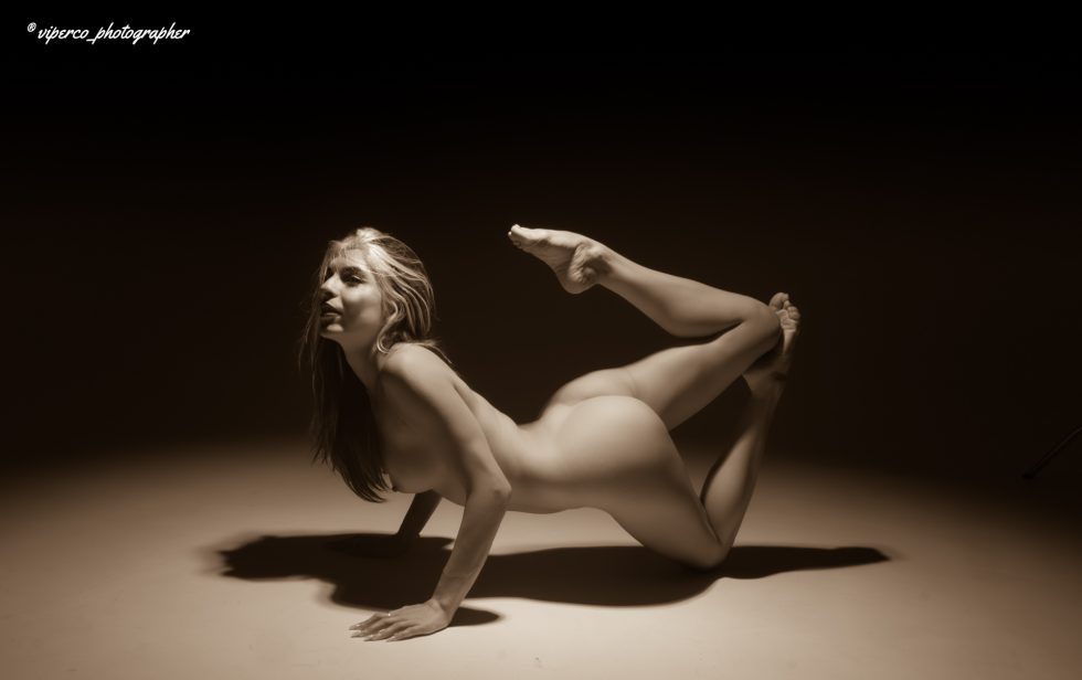 “Mujer arrodillada en un set de fotografía en blanco y negro, con el torso desnudo, cabeza inclinada hacia atrás y brazos abiertos, iluminada por un haz de luz circular que resalta su silueta.”