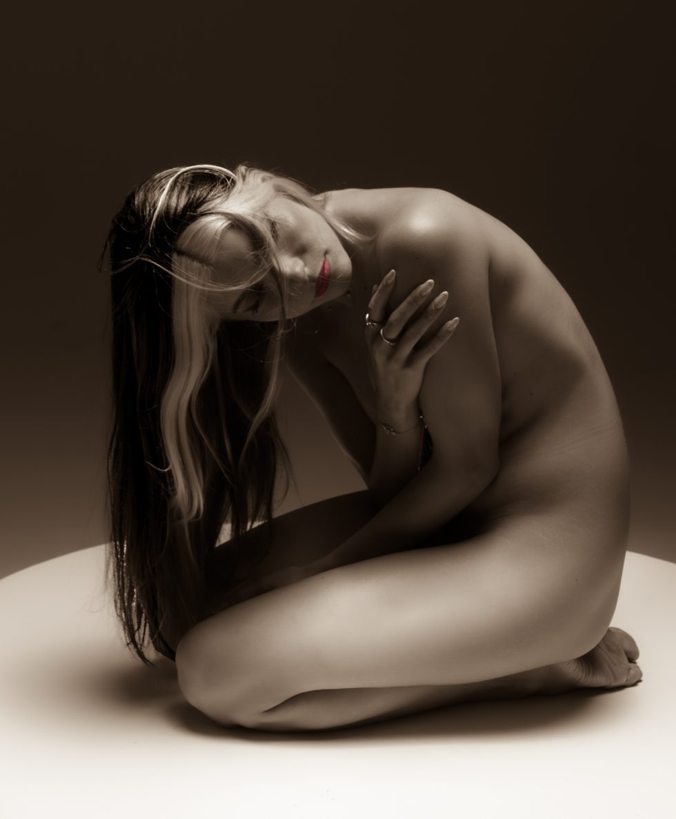 “Mujer arrodillada en un set de fotografía en blanco y negro, con el torso desnudo, cabeza inclinada hacia atrás y brazos abiertos, iluminada por un haz de luz circular que resalta su silueta.”
