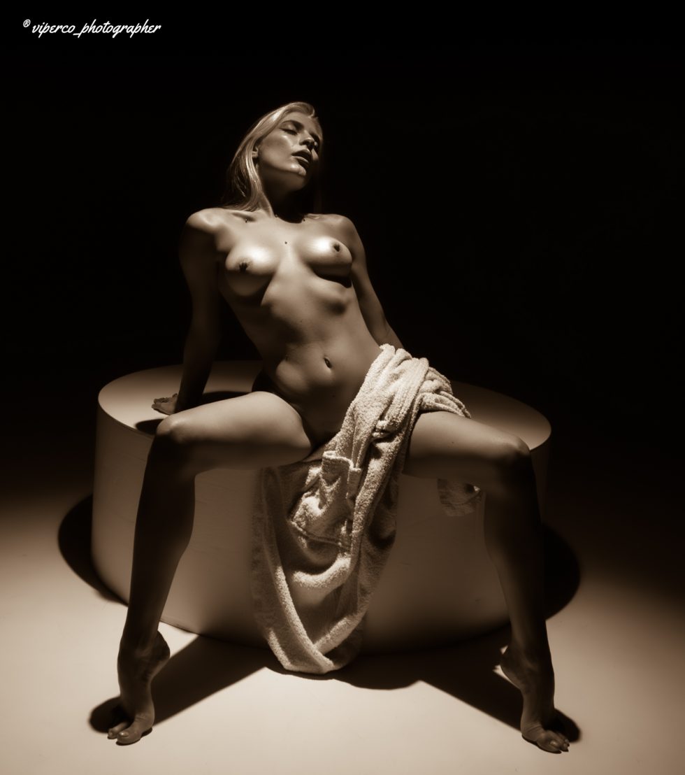 “Mujer arrodillada en un set de fotografía en blanco y negro, con el torso desnudo, cabeza inclinada hacia atrás y brazos abiertos, iluminada por un haz de luz circular que resalta su silueta.”